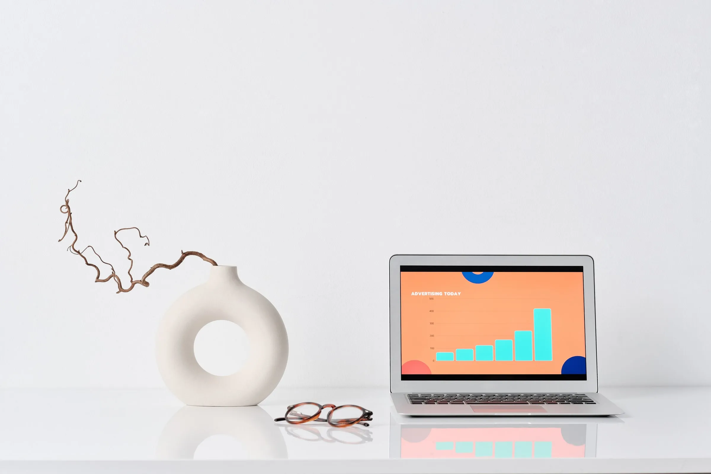 A laptop on a white desk showing a bar chart, beside a vase and a pair of glasses